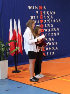 Dwie uczennice klasy siódmej stoją obok siebie trzymając w dłoniach rozłożone bordowe okładki. Dziewczynki patrzą na nie. za nimi widoczne są flagi Polskie.