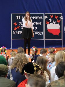 Uczennica klasy siódmej stoi na granatowym tle trzymając w dłoni przed sobą mikrofon. W tle za nią widoczna jest granatowa tablica z wyciętą mapą polski w kolorze biało czerwonym oraz przymocowanymi nad nią białymi i czerwonymi motylami.