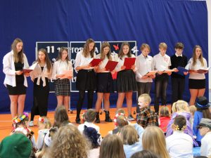 Uczniowie klasy siódmej stoją w rzędzie na scenie. Dzieci trzymają przed sobą bordowe podkładki, na które spoglądają. Przed nimi widoczne są głowy dzieci siedzących na materacach.