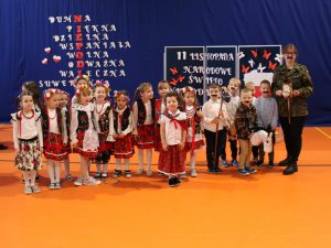 Wychowawczyni oddziału przedszkolnego w moro kurtce i przyklejonymi czarnymi wąsami trzyma przed sobą głowę pluszowego kucyka umieszczoną na patyku. Obok stoją obok siebie jej uczniowie. Dziewczynki ubrane są w stroje krakowianek z kolorowymi wiankami na głowie, a chłopcy mają przyczepione czarne wąsy i trzymają podobnie jak wychowawczyni głowy pluszowych koników umieszczonych na kijach.