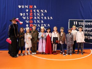 Wychowawczyni klasy drugiej w eleganckiej czarnej sukni i dużym kapeluszu na głowie pozuje do wspólnego zdjęcia wraz ze swoimi uczniami, którzy ubrani są w stroje postaci historycznych. Wszyscy stoją w jednym rzędzie na granatowym tle.