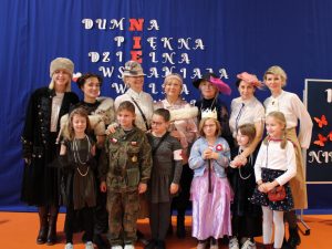 Uczniowie przebrani za postacie historyczne pozują do wspólnego zdjęcia wraz z nauczycielkami, które również ubrane są w stroje dam w kapeluszach, eleganckich bluzkach i długich spódnicach.