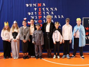 Nauczycielka w długim czarnym płaszczu oraz puchatej czapce na głowie pozuje do wspólnego zdjęcia z uczniami klasy V, którzy ubranie są w stroje postaci historycznych. Wszyscy stoją w jednym rzędzie.