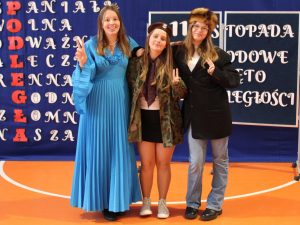 Trzy uczennice klasy siódmej pozuje do wspólnego zdjęcia stojąc obok siebie w swoich przebraniach. Wszystkie dziewczynki się uśmiechają i trzymają skierowane do góry dłonie z wyprostowanymi dwoma środkowymi palcami.