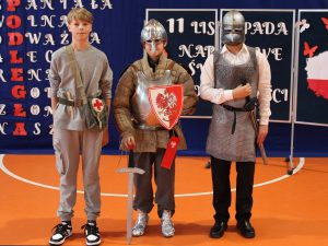 Trzech uczniów klasy siódmej pozuje do wspólnego zdjęcia. Dwoje z nich ma na sobie stroje rycerzy z hełmami na głowach, a trzeci chłopiec jest w szarym dresie z przepasana bokiem torbą ze znakiem krzyża naklejonym na klapie.