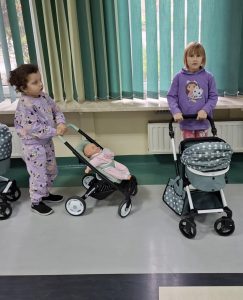 Dwie dziewczynki stoją na szarym pasie sali. Dziewczynki popychają przed sobą wózki dla lalek.