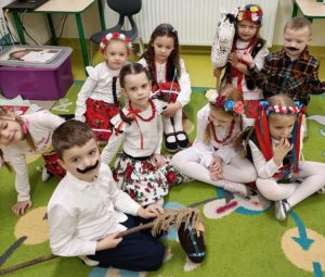 Uczniowie oddziału przedszkolnego siedzą na podłodze . Dziewczynki są ubrane w stroje krakowianek a chłopcy maja doklejone czarne wąsy. Dwoje dzieci trzyma w dłoniach drewniane patyki na których umieszczone są popiersia kucyków.