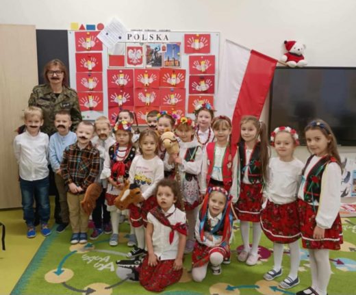 Uczniowie oddziału przedszkolnego pozują do wspólnego zdjęcia wraz ze swoją wychowawczynią. Wychowawczyni jest ubrana w żołnierski mundur i ma przyklejone czarne wąsy. Dziewczynki ubrane są w stroje krakowianek a chłopcy maja doklejone wąsy i trzymają w dłoniach przyczepione do drewnianych patyków pluszowy głowy kucyków. W tle za dziećmi wisi polska flaga oraz widoczna jest tablica z napisem "POLSKA:" raz przyczepionymi pracami plastycznymi uczniów przedstawiającymi polskie godło.