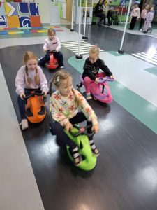 Czworo dzieci z oddziału przedszkolnego jedzie obok siebie po drodze wytyczonej na podłodze sali toruńskiego oddziału WORD. Dzieci siedzą na pojazdach z kółkami i się uśmiechają.
