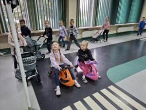 Dwie uczennice widoczne na pierwszym planie siedzą obok siebie na pojazdach z kółkami ustawionymi tuż przed przejściem dla pieszych w sali toruńskiego oddziału WORD. Dziewczynki patrzą w górę na sygnalizację umieszczoną przed przejściem, oczekując na zielone światło. Za nimi widoczni są uczniowie pchający przed sobą wózki dla lalek po ścieżce namalowanej wzdłuż zakręcającej do przejścia drogi.