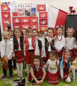 Uczniowie oddziału przedszkolnego pozują do wspólnego zdjęcia. dziewczynki ubrane są w stroje krakowianek a chłopcy maja doklejone wąsy i trzymają w dłoniach przyczepione do drewnianych patyków pluszowy głowy kucyków. W tle za dziećmi wisi polska flaga oraz widoczna jest tablica z napisem "POLSKA:" raz przyczepionymi pracami plastycznymi uczniów przedstawiającymi polskie godło.