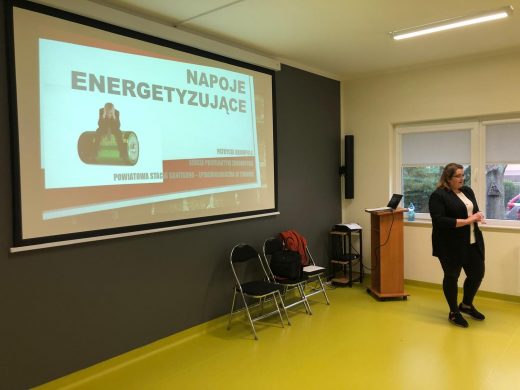 Na czarnej ścianie sali teatralnej szkoły rozwinięty jest ekran na którym widoczny jest slajd z prezentacji. Na ekranie widoczny jest duży napis "NAPOJE ENERGETYZUJĄCE", a pod nim grafika mężczyzny siedzące na dużej baterii opierającego głowę o swoje dłonie. Przed ekranem stoi kobieta prowadząca wykład.