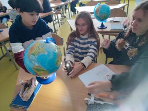 Troje uczniów siedzi przy wspólnym stole obok siebie. Chłopiec lekko podnosi nad stolik globus, który oświetlony jest przez światło latarki wychodzące z telefonu trzymanego przez uczennicę siedząca obok. Całej scenie przygląda się trzecia dziewczynka, patrząca w stronę globusa.