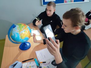Dwoje uczniów siedzi obok siebie przy stoliku, na którym ustawiony jest globus. Uczniowie trzymają w dłoniach telefony z włączonymi latarkami, których światło kierują w stronę globusa. Dziewczynka wolną dłoń zbliża do niego.