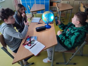 Troje uczniów siedzi przy stole, na którym ustawiony jest globus. Chłopcy siedzący na przeciwko siebie trzymają w dłoniach telefony z włączonymi latarkami, które kierują w stronę globusa. Siedząca obok dziewczynka przygląda się globusowi.
