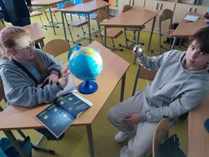 Dwoe uczniów siedzi przy stole, na którym ustawiony jest globus. Dziewczynka dotyka go palcami i patrzy w jego kierunku. Siedzący na przeciwko chłopak trzyma telefon z włączoną latarką skierowany na globus i patrzy w stronę obiektywu aparatu.