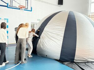 Uczniowie stoją w rzędzie w oczekiwaniu na wejście do nadmuchanej kopuły ustawionej na sali gimnastycznej. Jedna z uczennic wchodzi do środka. Kopuła jest ozdobiona srebrno granatowymi pasami.