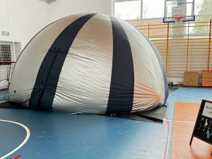 Zdjęcie kopuły mobilnego planetarium ustawionej na sali gimnastycznej. Kopuła jest ozdobiona srebrno granatowymi pasami.
