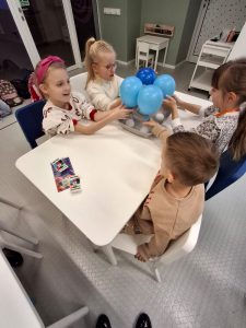 Czworo uczniów siedzi przy białym stoliku parami naprzeciwko siebie. Dzieci trzymają dłonie na niebieskich balonikach ustawionych na podstawce między nimi.