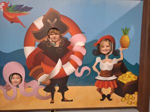 Trzy uczennice pozują do zdjęcia stojąc za makietą przez która widoczne są tylko ich głowy. Makieta przedstawia postać kobiety trzymającej w wyciągniętej w górę dłoni arbuza, pirata oraz ośmiornicy. Każda z postaci ma wycięty otwór na głowę, przez które wystają głowy uczennic.