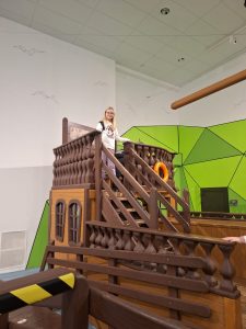 Uczennica stoi na szczycie rekonstrukcji statku. Opiera jedną dłoń o barierkę statku i patrzy wprost w stronę obiektywu.