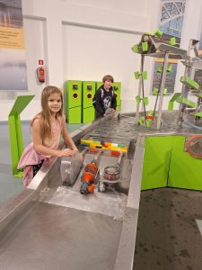 Dwoje uczniów stoi przy platformie imitującej rzekę, która jest wypełniona wodą. Dzieci patrzą w stronę obiektywu. Uczennica widoczna na pierwszym planie opiera dłonie o elementy mechaniczne tworzące śluzy w wybudowanej z klocków tamie na rzece.