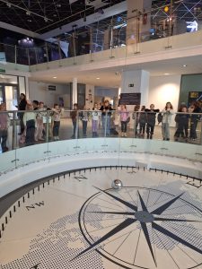 Uczniowie stoją na parterze Młyna Wiedzy wzdłuż przezroczystych barierek ustawionych w półokręgu. Dzieci patrzą na wahadło Fuko ustawione przed nimi z dużą srebrną kulą zawieszoną po środku.