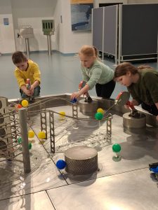 Troje uczniów pochyla się nad instalacją wykonaną z metalu, na której za pomocą dysz wody uczniowie przesuwają kolorowe piłeczki. Dzieci skupione są na ich obserwowaniu.