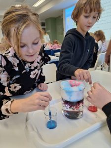 Dwoje uczniów pochyla się nad stołem, na którym ustawiona jest menzurka z kolorową cieczą i pianą u góry. Na pierwszym planie widoczna jest uczennica zaciągająca pipetą niebieski płyn ze zlewki stojącej przed nią. Jej kolega trzyma umieszczoną pipetę w zlewce zawierającej czerwony barwnik.