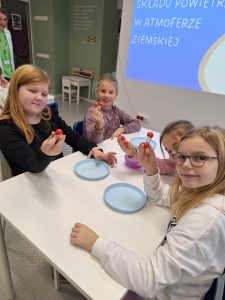 Cztery uczennice siedzą przy wspólnym białym stoliku na którym ustawione są plastikowe, niebieskie talerzyki. Wszystkie dziewczynki patrzą w stronę obiektywu, trzymając w dłoniach czerwone kuleczki połączone ze sobą metalowymi drucikami.