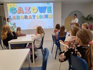 Uczniowie siedzą przy stolikach patrząc w stronę ekranu, na którym wyświetlony jest slajd z kolorowym napisem "GAZOWE LABOLATORIUM".