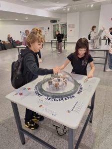 Dwoje uczniów stoi przy stoliku , z którego wystaje szklana kopuła, wokół której widoczny jest napis "AKCELERATOR". Dzieci trzymają dłonie umieszczone na szczycie kopuły.