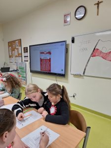 Uczennice edukacji wczesnoszkolnej siedzą przy stolikach i pochylają się nad krzyżówkami, które leżą przed nimi. W tle widać białą tablicę z narysowaną na niej odręcznie flagą Polski oraz dwoma napisami obok "JÓZEF", "HYMN". Obok na ekranie wyświetlony jest slajd z białym tekstem umieszczonym na czerwonym tle, nad którymi widnieje polska flaga i nasze godło narodowe.