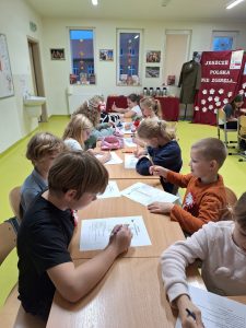 Uczniowie oddziału wczesnoszkolnego siedzą przy długim stole. Dzieci pochylają się nad krzyżówkami, leżącymi przed nimi. W tle widać przygotowaną na spotkanie dekorację - na czerwonym materiale umieszczony jest biały napis "JESZCZE POLSKA NIE ZGINĘŁA..." a pod nim białe kwiaty. Obok na manekinie zawieszony jest długi płaszcz.
