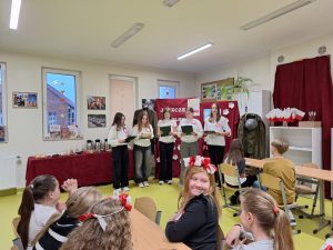 Pięć uczennic starszych klas stoi obok siebie trzymając przed sobą podkładki, nad którymi pochylają głowy. dziewczynki mają przyczepione biało-czerwone kotyliony. Przed nimi widać głowy uczestniczących w spotkaniu uczniów z oddziału wczesnoszkolnego, którzy siedzą na krzesełkach. Za dziewczynkami ustawiona jest czerwona dekoracja.