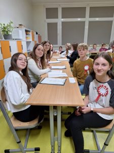 Uczniowie szkolnego wolontariatu biorący udział w spotkaniu siedzą przy długim stole. Dzieci patrzą w stronę obiektywu a przed nimi leżą podkładki z białymi kartkami. W tle widać siedzących w dalszej części sali uczniów oddziału wczesnoszkolnego.