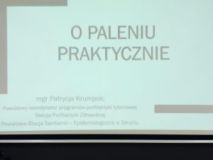 Zdjęcie wyświetlanego na rzutniku slajdu. Na białym tle z czarną, częściowo otwartą ramką widoczne są napisy: "O PALENIU PRAKTYCZNIE", "mgr Patrycja Krumpolc Powiatowy koordynator programów profilaktyki tytoniowej Sekcja Profilaktyki Zdrowotnej Powiatowa Stacja Sanitarno-Epidemiologiczna w Toruniu".