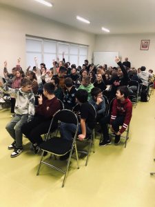 Uczniowie siedzą na krzesłach ustawionych na sali teatralnej. Część z nich podnosi wysoko w górę rękę zgłaszając się. Dwóch chłopców siedzących z przodu odwraca się za siebie.