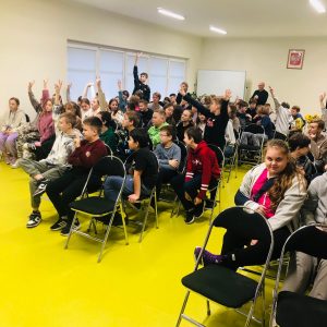 Uczniowie siedzą na krzesłach ustawionych na sali teatralnej. Część z nich podnosi wysoko w górę rękę zgłaszając się.