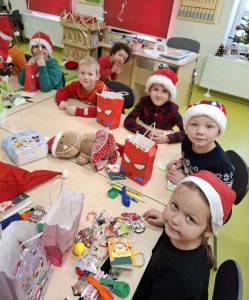 Uczniowie oddziału przedszkolnego siedzą przy dużym stole. Przed dziećmi ustawione są papierowe torby z otrzymanymi upominkami. Część z prezentów leży przed dziećmi na stole. Wszyscy uczniowie patrzą w stronę obiektywu i się uśmiechają.