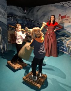 Oddział przedszkolny w "domu Legend". Dwie uczennice stoją na przeciwko siebie na drewnianych podkładkach ustawionych na krótkich belkach. Dziewczynki trzymają tyczki na końcach których zawieszone są worki wypełnione pierzem. Z tyłu stoi kobieta podpierająca swoje dłonie o biodra.