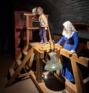 Oddział przedszkolny w "domu Legend". Uczennica stoi na drewnianej platformie. Obok opiera się o jedną z belek platformy kobieta w granatowej sukni i białej chuście na głowie. Przed kobietą wisi dzwon.
