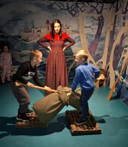 Oddział przedszkolny w "domu Legend". Dwóch chłopców stoi na przeciwko siebie na drewnianych podkładkach ustawionych na krótkich belkach. Uczniowie trzymają tyczki na końcach których zawieszone są worki wypełnione pierzem, które krzyżują przed sobą. Z tyłu stoi kobieta podpierająca swoje dłonie o biodra.