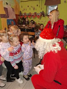 Uczniowie oddziału przedszkolnego stoją w kolejce przed Świętym Mikołajem siedzącym przed nimi. Dziewczynka stojąca przed nimi podnosi w górę ręce z wyciągniętymi palcami.