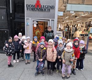 Uczniowie wraz ze swoją wychowawczynią pozują do wspólnego zdjęcia wraz ze swoją wychowawczynią stojąc przed wejściem do "DOMU LEGEND TORUŃSKICH".