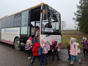 Uczniowie oddziału przedszkolnego wchodzą do autobusu, który za przednią szybą ma tabliczkę z napisem "OSTASZEWO SZKOŁA".