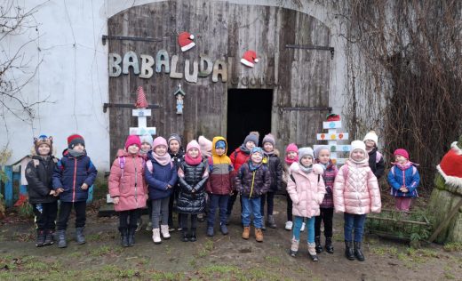 Uczniowie oddziału przedszkolnego pozują do wspólnego zdjęcia stojąc przed budynkiem z otwartymi drzwiami, na którym umieszczony jest napis "BABALUDA" oraz dwie mikołajkowe czapki.