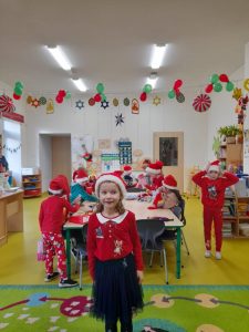 Na pierwszym planie widoczna jest uczennica stojąca na zielonym dywanie. Dziewczynka uśmiecha się a na głowie ma czerwoną czapeczkę. Za nią przy stole siedzą pozostali uczniowie. Nad nimi rozciągnięta jest girlanda tworząca świąteczną dekorację.