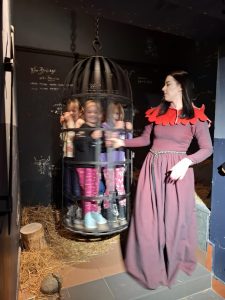 Oddział przedszkolny w "domu Legend". Uczniowie sa zamknięci w klatce zawieszonej tuz nad ziemią. Obok klatki stoi kobieta w bordowej sukni, która nią obraca. Pani patrzy w kierunku uczniów.