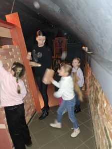 Oddział przedszkolny w "domu Legend". Uczennice ustawiają mur z cegieł Jedna z nich stoi przy murze a druga przed nim trzymając w ręku cegłę. Obok muru stoi nauczycielka patrząca na swoją podopieczna, która trzyma cegłę. Sylwetka dziewczynki jest rozmazana.